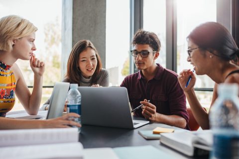 Giovani di diverse etnie studiano per l'università in gruppo seduti attorno a un tavolo. Studenti universitari riuniti con libri e laptop per cercare informazioni per il loro progetto